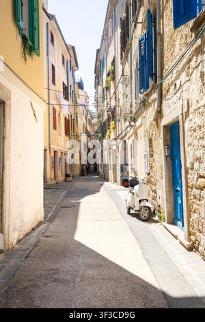 Piran, Slowenien. 26. Mai 2026. Eine schmale, sonnendurchflutete Gasse, gesäumt von hohen, gelben Gebäuden. Die Straße ist mit Stein und Asphalt gepflastert, und über Kopf kreuzen sich Stromleitungen. Stockfoto