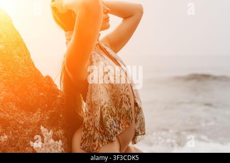 Strandfrau Sonnenuntergang junge Frau trägt ein gemustertes Cover-up steht gegen einen Felsen am Ozean zur Goldenen Stunde Stockfoto