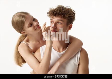 Liebevolle junge Frau umarmt ihren Freund, während sie Creme unter den Augen aufträgt Stockfoto