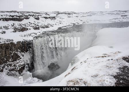 Der mächtige isländische Dettifoss-Wasserfall fließt kraftvoll zwischen felsigen Schluchten und völlig schneebedeckten Klippen. Ein dichter Wasserdunst steigt auf von Stockfoto