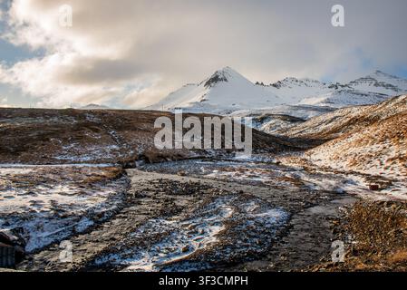 Ein felsiger Bach windet sich durch ein teilweise schneebedecktes Hügelland in Island. Majestätische schneebedeckte Berge erheben sich im Hintergrund, während der Sk Stockfoto