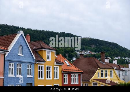 Lebendige Reihe farbenfroher Holzhäuser in Bergen, Norwegen, mit einem üppigen bewaldeten Hügel und einer Seilbahn unter bewölktem Himmel. Stockfoto