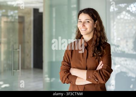 Professionelle Frau mit einem warmen Lächeln und verschränkten Armen, die selbstbewusst in einem hellen zeitgenössischen Büro steht und Führung und Erfolg in einem Unternehmensumfeld repräsentiert Stockfoto