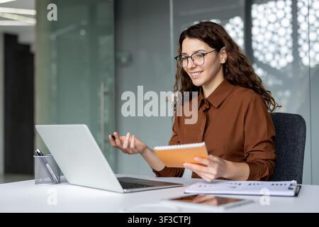 Lächelnde junge Geschäftsfrau mit Brille, die an einem modernen Bürotisch an einer virtuellen Videokonferenz auf ihrem Laptop teilnimmt, aktiv kommuniziert und während des Online-Meetings Notizen macht Stockfoto