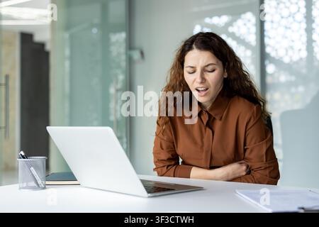 Frau mit Magenschmerzen und Krämpfen, die am Schreibtisch mit einem Laptop sitzt, betont Arbeitsplatzgesundheitsprobleme, Stress und medizinisches Unbehagen, die die Produktivität beeinträchtigen Stockfoto