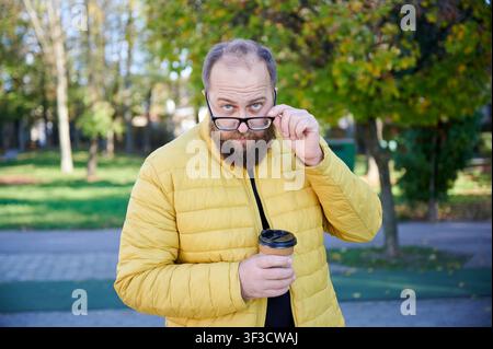 Bärtiger Mann senkt die Brille und schaut in die Kamera, während er im Park einen To-go-Kaffee hält. Ideal für Lifestyle, lässige Mode, Herbst, Augenpflege oder urbane Werbung Stockfoto