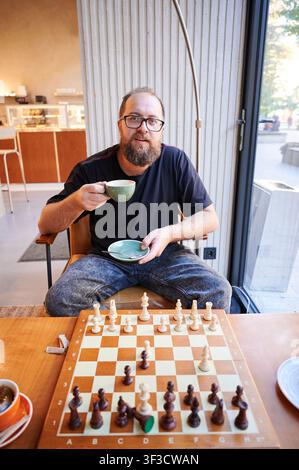 Mann, der der Kamera gegenübersteht, mit Schachbrett und Tasse, einladender Ausdruck und Bereitschaft für einen Rückkampf, lässige Spielumgebung mit Glasfensterblick, Gebäck auf dem Teller und warmes Umgebungslicht, gesprächige Stimmung Stockfoto