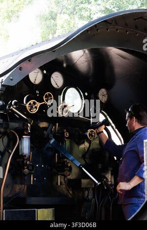 Swanage, England, 18. Juli 2026. Ingenieur bedient die Steuerungen einer Dampflokomotive, Messgeräte mit SR sichtbar in der Kabine, Dampf steigt draußen gegen grüne Bäume auf Stockfoto