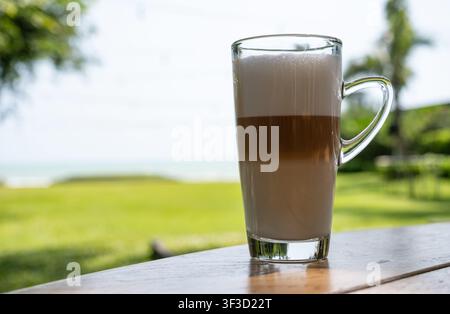 Schichtweiser Latte-Kaffee in einer klaren Glastasse auf einem Holztisch mit verschwommenem grünem Gras und Seeblick Stockfoto