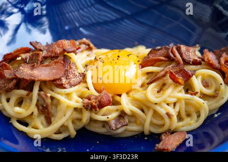 Spaghetti Carbonara mit knusprigem Speck und einem rohen Eigelb obendrauf, serviert auf einem blauen strukturierten Teller Stockfoto