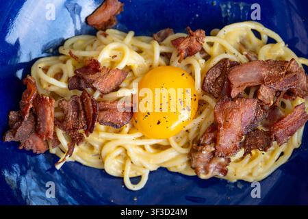 Oberblick auf Spaghetti Carbonara mit knusprigem Speck und einem rohen Eigelb obendrauf, serviert auf einem blauen strukturierten Teller Stockfoto
