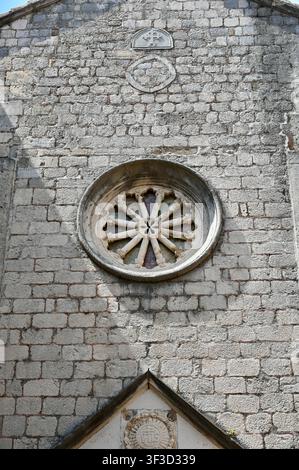 Nahaufnahme einer mittelalterlichen Steinkirchenfassade mit einem kunstvoll geschnitzten Rosettenfenster, Kreuzmotiv und alter Mauerarbeit, repräsentativ für traditionelle europäische Sakralarchitektur Stockfoto