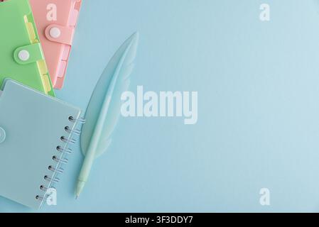 Hellgrüne, rosa und blaue Plastiknotizbücher zusammen mit einem hellblauen federförmigen Stift liegen ordentlich auf einer pastellblauen Oberfläche angeordnet, was sch repräsentiert Stockfoto