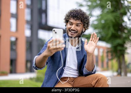 Lächelnder junger Mann mit lockigem Haar und Kopfhörern führt ein lebhaftes Videogespräch auf seinem Handy im Freien, winkt der Person auf dem Bildschirm zur Begrüßung zu und genießt die drahtlose Kommunikation Stockfoto