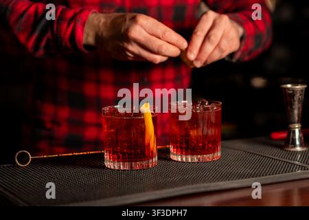 Barkeeper in kariertem Hemd bereitet zwei rote Cocktails in Longdrinkgläsern auf dunkler Holztheke vor. Warme bernsteinfarbene Abendstimmung. Ähnliche Cocktails: Neg Stockfoto