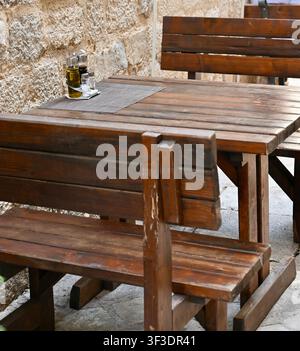 Gemütliche Außenterrasse eines Straßencafés mit rustikalem Holztisch, Holzbank und Gewürzetui in der Nähe einer alten Steinmauer im Hintergrund. Ideales Konzept für europäisches Essen, Reisen, Sommertourismus und Gastronomie. Stockfoto