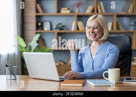 Erfahrene Geschäftsfrau fühlt sich aufgeregt und glücklich, nachdem sie ein wichtiges Geschäftsziel erreicht oder gute Nachrichten erhalten hat, während sie aus der Ferne an ihrem Laptop in einem Home-Office-Umfeld arbeitet Stockfoto