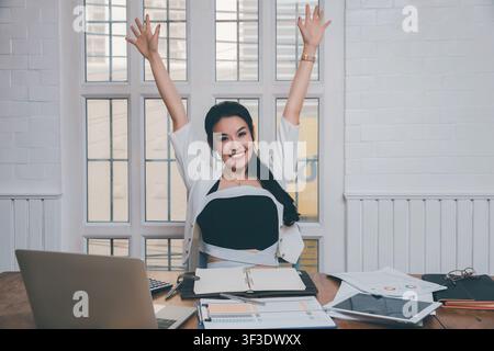 Zufriedene junge asiatische Geschäftsfrau sitzt mit Laptop und erhobener Hand. Überglückliche motivierte Frau fühlt sich wie eine Gewinnerin und freut sich über den Online-Gewinn einer neuen Jobchance Stockfoto