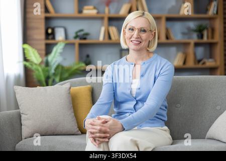 Seniorin mit Brille und blauem Cardigan, die selbstbewusst lächelt, während sie drinnen auf einem bequemen Sofa sitzt vor dem Hintergrund eines modernen Wohnzimmerschranks, verkörpernd Entspannung und Anmut Stockfoto