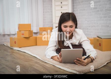 Geschäftsfrau nimmt Bestellungen von Kunden über Online-Verkäufe entgegen, während sie auf einem Teppich in ihrem Heimbüro liegt, bereitet eine Paketbox mit Produkten für die Lieferung vor Stockfoto