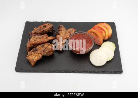 Gegrillte Schweinerippchen serviert mit Tomatensoße, geschnittenen Tomaten und Zwiebel auf einem schwarzen Schieferbrett als frisch zubereitetes Barbecue-Gericht. Stockfoto