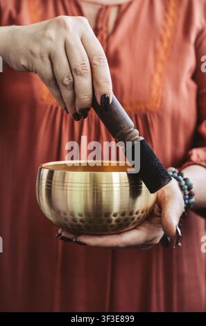 Nahaufnahme von weiblichen Händen, die eine Messing-Sing-Schale halten, in einem fließenden Terrakotta-Kleid und mit Perlenarmband. Konzept von Achtsamkeit, Entspannung, Spi Stockfoto