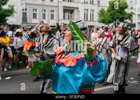 Berlin, Deutschland - 24. Mai 2026. Eine lächelnde Darstellerin in einem türkisfarbenen und orangefarbenen Kostüm tanzt mit grünen Blättern während des Karneval der Kulturen auf der Straße Stockfoto