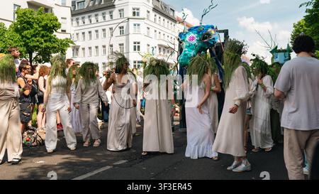 Berlin, Deutschland – 24. Mai 2026. Darsteller in weißer Kleidung und Gras-Kopfschmuck paradieren neben einer riesigen türkisfarbenen Drachenpuppe während des Karnevals der K Stockfoto
