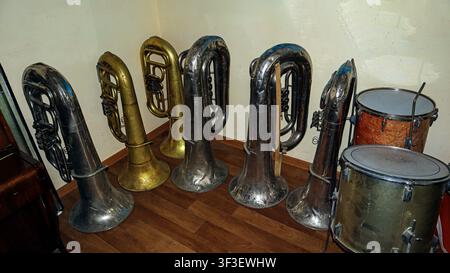 Alte Messinginstrumente, darunter Tuben und eine Trommel, sind ordentlich in einem Lagerraum der Musikschule angeordnet und zeigen die Geschichte der musikalischen Bildung. Stockfoto