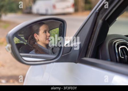 Junge Frau im Seitenrückspiegel eines Autos sichtbar, während sie fährt, Außenansicht, geparkte Autos und Bäume im Hintergrund, Konzept für Verkehrssicherheit und Fahren Stockfoto