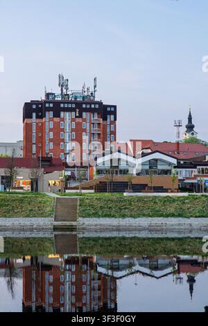 Vinkovci, Kroatien - 15. April 2026: Flusspromenade mit einer Mischung aus historischen und modernen Gebäuden, die sich im ruhigen Flusswasser spiegeln, grasbewachsener Flussufer und Stockfoto
