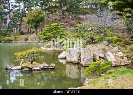 Eine ruhige japanische Gartenlandschaft mit einem ruhigen Teich mit grünlichem Wasser, das die umgebende Landschaft widerspiegelt. Mehrere kleine Inseln übersäen den Teich Stockfoto