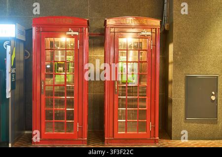 Zwei traditionelle rote Telefonzellen im britischen Stil stehen nebeneinander in einem Raum an einer braunen Fliesenwand. Beide Kabinen tragen das Wort "TELEFON" in Stockfoto