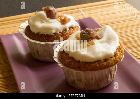 Zwei Kürbismuffins in Papierbechern mit weißer Glasur und kandierter Kastanie auf einem lila Teller, Bambusmatte als Hintergrund, 45-Grad-Ansicht Stockfoto