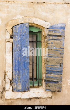 Eine Nahaufnahme zeigt Details eines rustikalen Fensters, das von verwitterten blauen Holzläden auf einer Steinmauer in Arles, Frankreich, eingerahmt ist. Stockfoto
