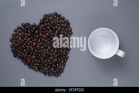 Kaffeebohnen in Herzform mit leerer Tasse auf grauem Hintergrund Stockfoto