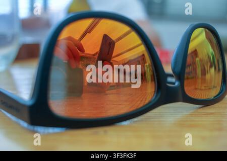 Nahaufnahme von schwarzen Sonnenbrillen auf einem Holztisch, die eine scharfe orangefarbene Reflexion einer Person zeigen, die drinnen ein Mobiltelefon hält. Stockfoto