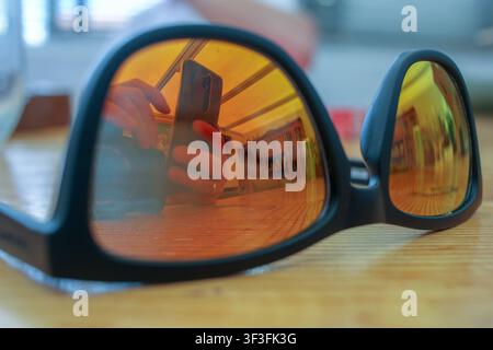 Eine Detailansicht von modernen Sonnenbrillen, die auf einer Oberfläche liegen, mit einer klaren Reflexion eines Smartphone-Nutzers, der eine Innenszene aufnimmt. Stockfoto
