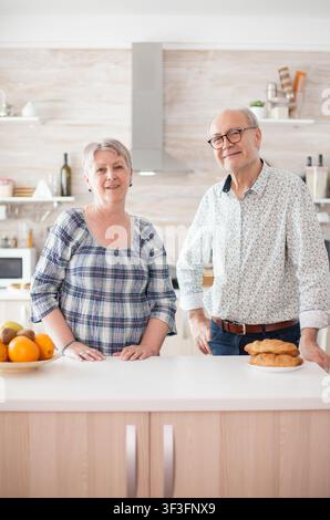 Seniorenpaar lächelt in einer Hausküche in die Kamera und zeigt einen fröhlichen Ruhestandsstil, Zuneigung und entspannte häusliche Gemütlichkeit. Stockfoto