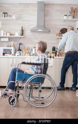 Senior entspackt Einkäufe in der Küche, während seine behinderte Frau im Rollstuhl sitzt, was häusliche Unterstützung und fürsorgliche Partnerschaft zeigt, und Frühstück, Rollstuhl und das sichtbare Zuhause ausbalanciert. Stockfoto