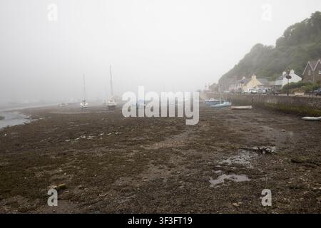 Boote und Yachten, die bei Ebbe auf dem schlammigen Meeresboden in einem Küstenhafen gestrandet sind, eingehüllt in dichten Meeresnebel und Dunst. Stockfoto