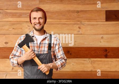 Lächelnder junger Holzfäller oder Zimmermann in kariertem Hemd und Beanie, der eine Axt vor einer hölzernen Wand hält, Er steht selbstbewusst gegen ein rusti Stockfoto