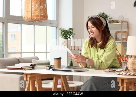 Lächelnde junge Frau liest Dokumente an einem modernen Schreibtisch im Heimbüro mit Kaffee und Designmaterialien Stockfoto