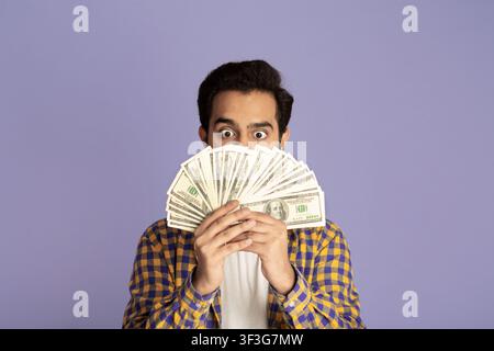 Gewinnen Sie Lotterie. Aufgeregt indischen Kerl mit Haufen Geld über violetten Hintergrund Stockfoto