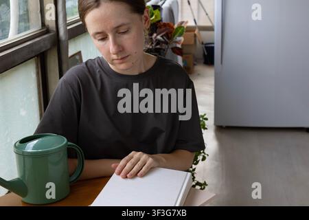 Junge Frau liest ein leeres Hardcover-Buch zu Hause mit einem grünen Gießkanne Stockfoto