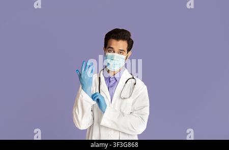Schutz vor Viruserkrankungen. Indischer Arzt trägt medizinische Maske, Laborkittel und Gummihandschuhe auf violettem Hintergrund Stockfoto