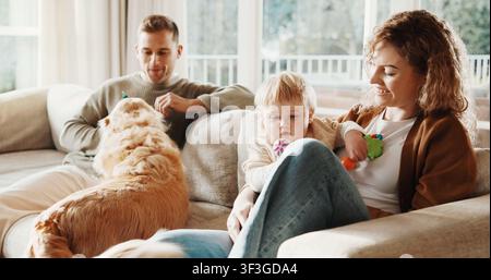 Eltern, Kind und Hund mit Liebe auf der Couch für Fürsorge, Spiel und Lächeln für Bindung oder Verbindung zu Hause. Vater, Mutter und Baby im Wohnzimmer für das Haustier Stockfoto