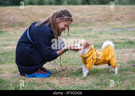 Frau mit Fütterung ihres süßen Hundes, der einen gelben Regenmantel trägt, im Park. Haustierbesitzerin spielt mit dem Hund im Freien. Freundschaft zwischen Mensch und Tier. Fürsorge und Stockfoto