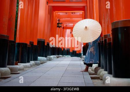 Niedrige Seitenansicht einer Frau in blauem Kimono mit Papier-Regenschirm, die in einem roten Torii-Tunnel am Fushimi Inari steht Stockfoto