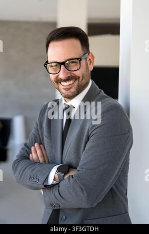 Junger glücklicher, erfolgreicher Geschäftsmann mit überkreuzten Armen lächelnd im Firmenbüro Stockfoto